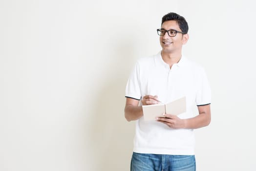 Portrait of Indian guy taking note on book, looking at side and smiling. Asian man standing on plain background with shadow and copy space. Handsome male model.