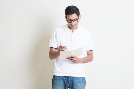Portrait of Indian guy taking note on book. Asian man standing on plain background with shadow and copy space. Handsome male model.