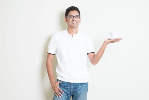 Indian guy holding a cup of coffee on hand. Asian man standing on plain background with shadow and copy space. Handsome male model.