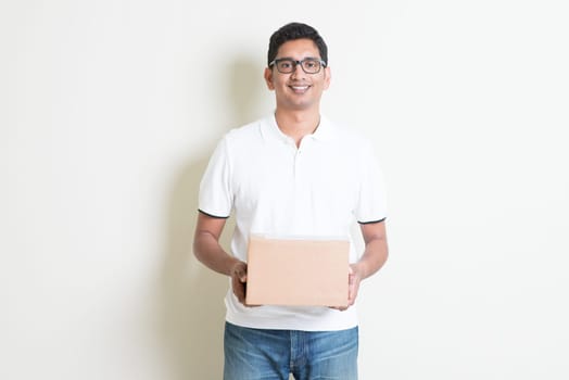 Indian man smiling and holding a courier box standing on plain background with shadow. Asian handsome guy model.