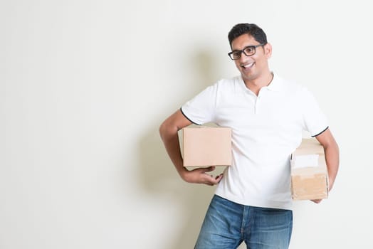 Courier delivery service concept. Happy Indian man holding brown boxes, standing on plain background with shadow. Asian handsome guy model.