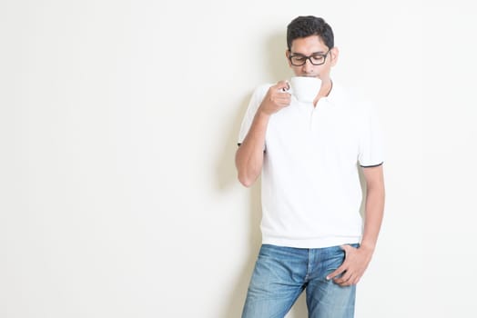 Indian guy drinking coffee. Asian standing on plain background with shadow and copy space. Handsome male model.