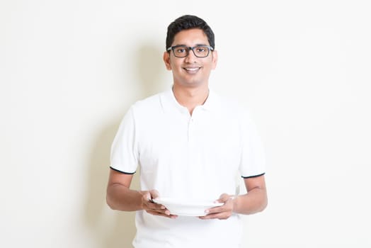 Dining concept. Indian chef holding an empty plate on hand, ready for food. Asian man standing on plain background with shadow and copy space. Handsome male model.