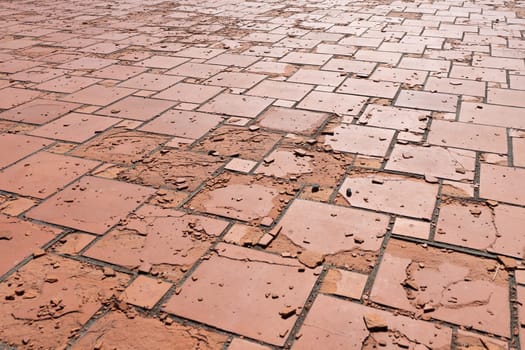 Red square floor tiles damaged by weathering or material defects. The surface is crumbling and the plates are disintegrating.