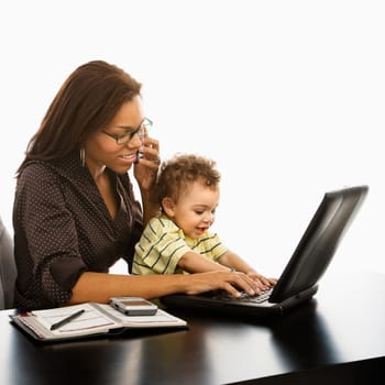 African American businesswoman at work on laptop and cell phone with toddler son on lap.