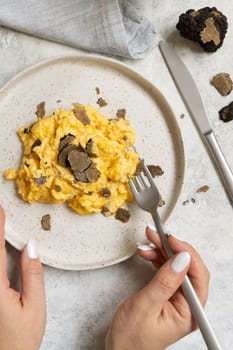Hand with fork over plate with Scrambled eggs with fresh black truffles top view. Italian cousine, gourmet breakfast