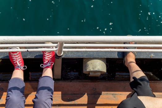 Top view showing legs of two persons, sitting on a narrow sea side passage of a ferry boat, looking toward the sea, and putting their legs on passage handrail
