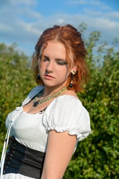Portrait of a pretty young woman wearing white top, looking down, standing outdoors with green trees and blue sky