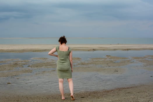 Young woman in a short green dress is standing in front of the sea on a sandy beach