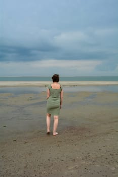 Young woman in a short green dress is standing in front of the sea on a sandy beach