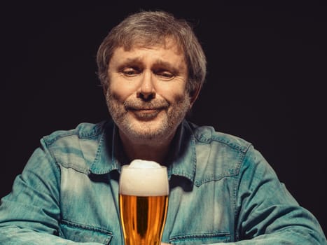 Enjoying his favorite beer.  The front view of handsome spellbound man in denim shirt with glass of beer, sitting at the wooden table. Concept of desire and love for beer and tender emotion