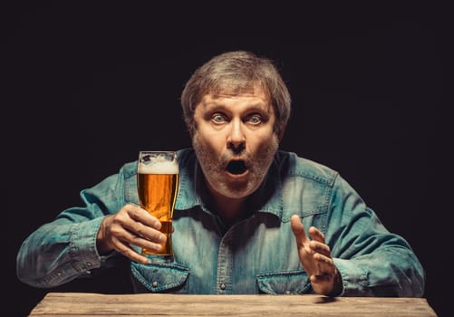 The front view of handsome man as enchanted and emotional fan in denim shirt with glass of beer, sitting at the wooden table. Concept of  surprise and exciting spectacle