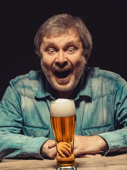 Enjoying his favorite beer.  The front view of handsome screaming man as fan in denim shirt with glass of beer, sitting at the wooden table. Concept of enthusiasm and ecstasy