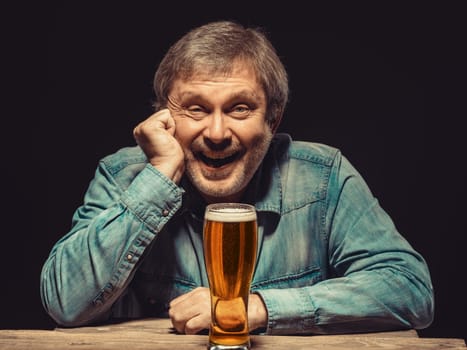 Enjoying his favorite beer.  The front view of handsome smiling  man as fan in denim shirt with glass of beer, sitting at the wooden table