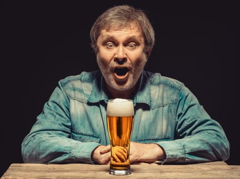 Enjoying his favorite beer.  The front view of handsome screaming man as fan in denim shirt with glass of beer, sitting at the wooden table. Concept of enthusiasm and ecstasy