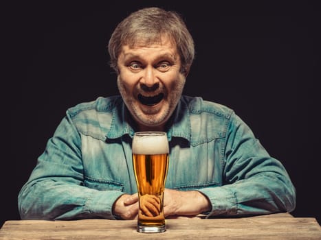 Enjoying his favorite beer.  The front view of handsome smiling  man as fan in denim shirt with glass of beer, sitting at the wooden table. Concept of enthusiasm and ecstasy
