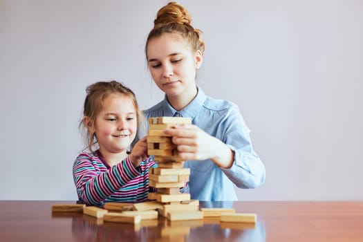 Little girl preschooler and her elder sister playing together with wooden blocks game toy. Siblings spending time sitting at desk at home