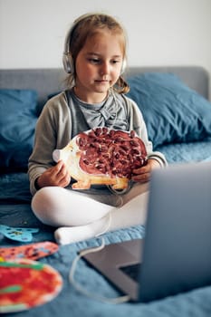 Little girl preschooler learning online showing her works drawings done at home. Child learning watching lesson remotely, talking with tutoress on video call from home during quarantine. Kid using laptop, headphones sitting on bed
