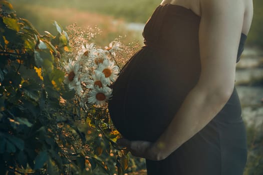 Pregnant woman hugging her tummy standing outdoors surrounded by nature. Pregnancy, expectation, motherhood concept