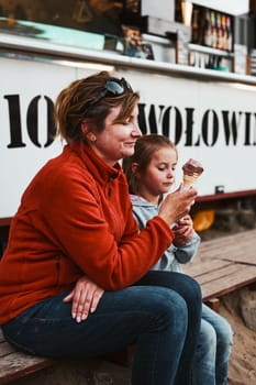 Mother and her daughter eating ice cream sitting on a step in front of food truck during summer vacations