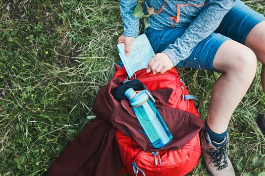 Woman with backpack having break during trip in mountains taking map out sitting on grass on summer vacation day