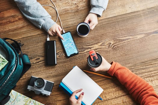 Family planning vacation trip. Girl searching travel destination and routes using navigation map on mobile phone. Woman writing journal and making notes. Charging smartphone with power bank. Using technology while travelling. People sitting at wooden table with camera and backpack during journey. Travel plan concept