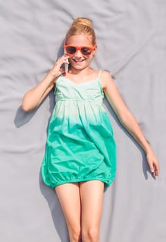 Young girl talking to her schoolfriend using smart phone, lying on plain blanket outdoors, wearing sunglasses