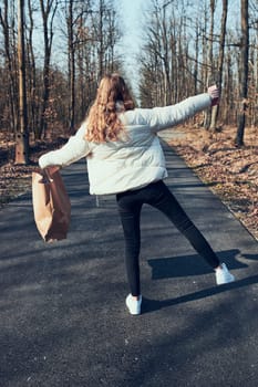 Young woman carrying big paper shopping bag. Back view of happy girl walking in a park after shopping, wearing white jacket and shoes. Real people, authentic situations