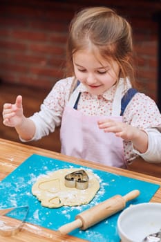 Little girl cutting the dough to heart shapes cookies. Kid taking part in baking workshop. Baking classes for children, aspiring little chefs. Learning to cook. Combining and stirring prepared ingredients. Real people, authentic situations