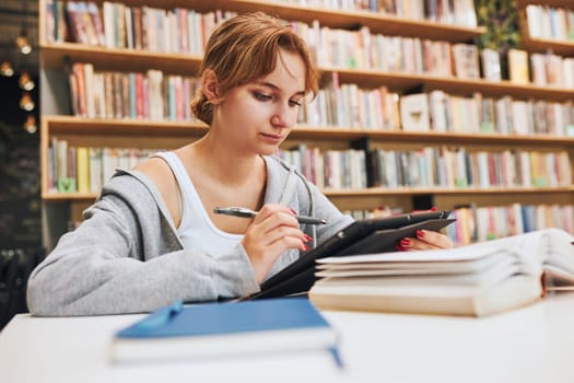 Student learning in university library. Young woman writing essay and making notes using computer tablet. Focused student studying for college exams. Using technology. Back to school