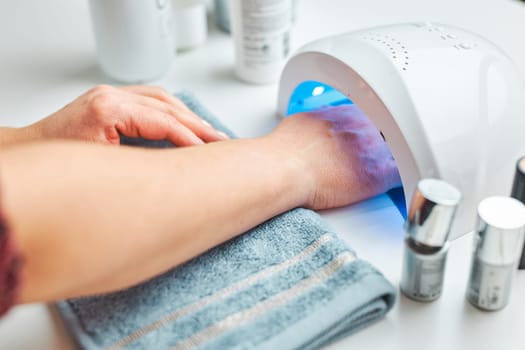 Woman applying gel hybrid polish using UV lamp. Beauty wellness spa treatment concept. Cosmetic products, UV lamp, green leaves on white table. Spa, manicure, skin care concept. Flat lay, overhead view