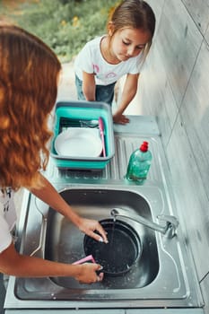 Teenager girl washing up the dishes pots and plates with help her younger sister in the outdoor kitchen during vacations on camping. Camp life. Sisters spending time and working together