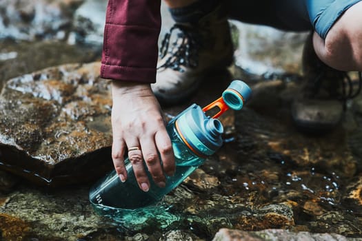 Woman taking pure water to bottle from mountain stream during trekking in mountains. Hiker crouching on rocks, filling bottle up with cold mountain water. Enjoying the outdoors in the summer trip vacation