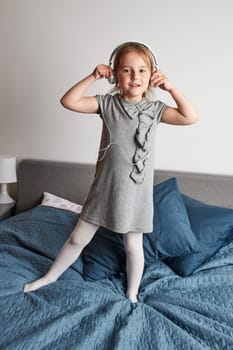 Little girl singing holding headphones cord imitating herself a real singer. Child having fun jumping dancing listening to music on bed in bedroom at home