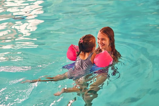 Young woman teaching to swim her younger sister and playing with her in swimming pool. Candid people, real moments, authentic situations