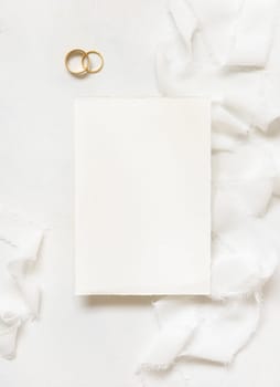 Blank card near white silk ribbons and wedding rings top view, wedding mockup. Romantic scene with vertical card and pastel flowers flat lay. Valentines, Spring or Mothers day concept