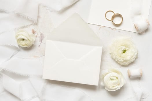 Blank envelope near cream roses, white silk ribbons and wedding rings top view, wedding mockup. Romantic scene with pastel flowers flat lay. Valentines, Spring or Mothers day concept