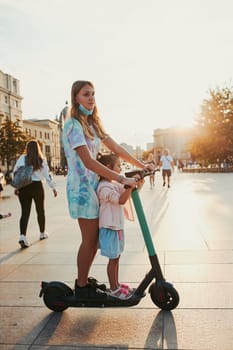 Teenager girl and her sister preschooler riding an electric scooter in the city center. Concept of happy family. Candid people, real moments, authentic situation