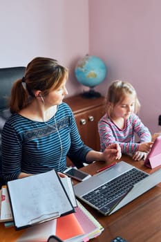 Woman mother working doing her job remotely during video chat call stream online course webinar on laptop from home while her daughter watching video on tablet. Woman sitting at desk in front of computer looking at screen