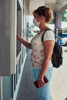 Woman withdrawing money from atm machine using debit or credit card standing outdoors in the street, wearing face mask to cover mouth and nose during pandemic coronavirus outbreak