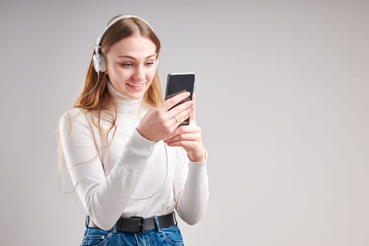 Young woman girl listening to music streaming content having fun watching video enjoying video chat talking with friends making gestures faces using smartphone earphones headphones standing over plain grey background