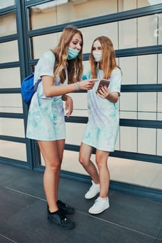 Young women girls using smartphones using social media sending texts watching photos and videos. Friends spending time talking with friends standing at store front downtown. Girls wearing the face masks to avoid virus infection and to prevent the spread of disease in time of coronavirus