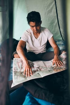 Young man looking at a map of mountain trails planning next trip on summer vacation sitting in a tent on camping. Concept of camp life