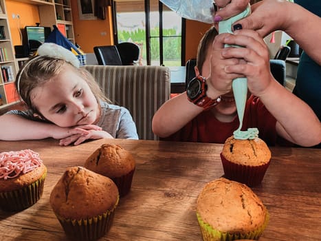 Group of children baking cupcakes, squeezing cream from confectionery bag, preparing ingredients, topping, sprinkles for decorating cookies. Kids cooking, working together in kitchen at home. Concept of happy family