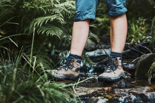 Female legs with trekking boots standing on stones in mountain stream. Summer vacation close to nature. Concept of actively spending leisure time