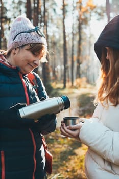 Mother and daughter with backpack having break during autumn trip pouring a hot drink from thermos flask on autumn cold day. Active women wandering in a forest actively spending time