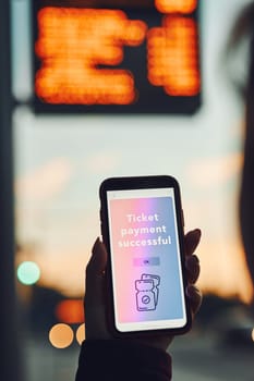 Person buying ticket online near bus stop before ride. Person paying for ticket using mobile payment app. Using technology on the go. Order confirmation on mobile screen. Top up account in app. Bus routes schedule in background