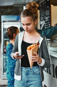 Teenage girl eating potato fries from carton cone standing in front of food truck. Teenager having a fast food meal outdoors during summer vacations