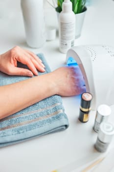 Woman applying gel hybrid polish using UV lamp. Beauty wellness spa treatment concept. Cosmetic products, UV lamp, green leaves on white table. Spa, manicure, skin care concept. Flat lay, overhead view