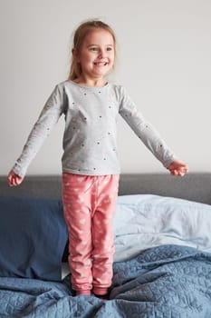 Little happy playful kid girl having fun making silly funny faces jumping on bed in bedroom in the morning at home